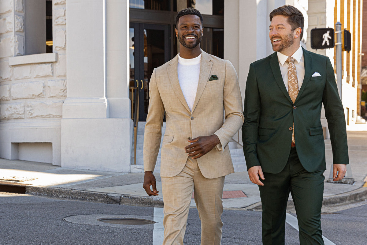 Two models walking across a downtown street, one in a tan suit and one in a dark green suit