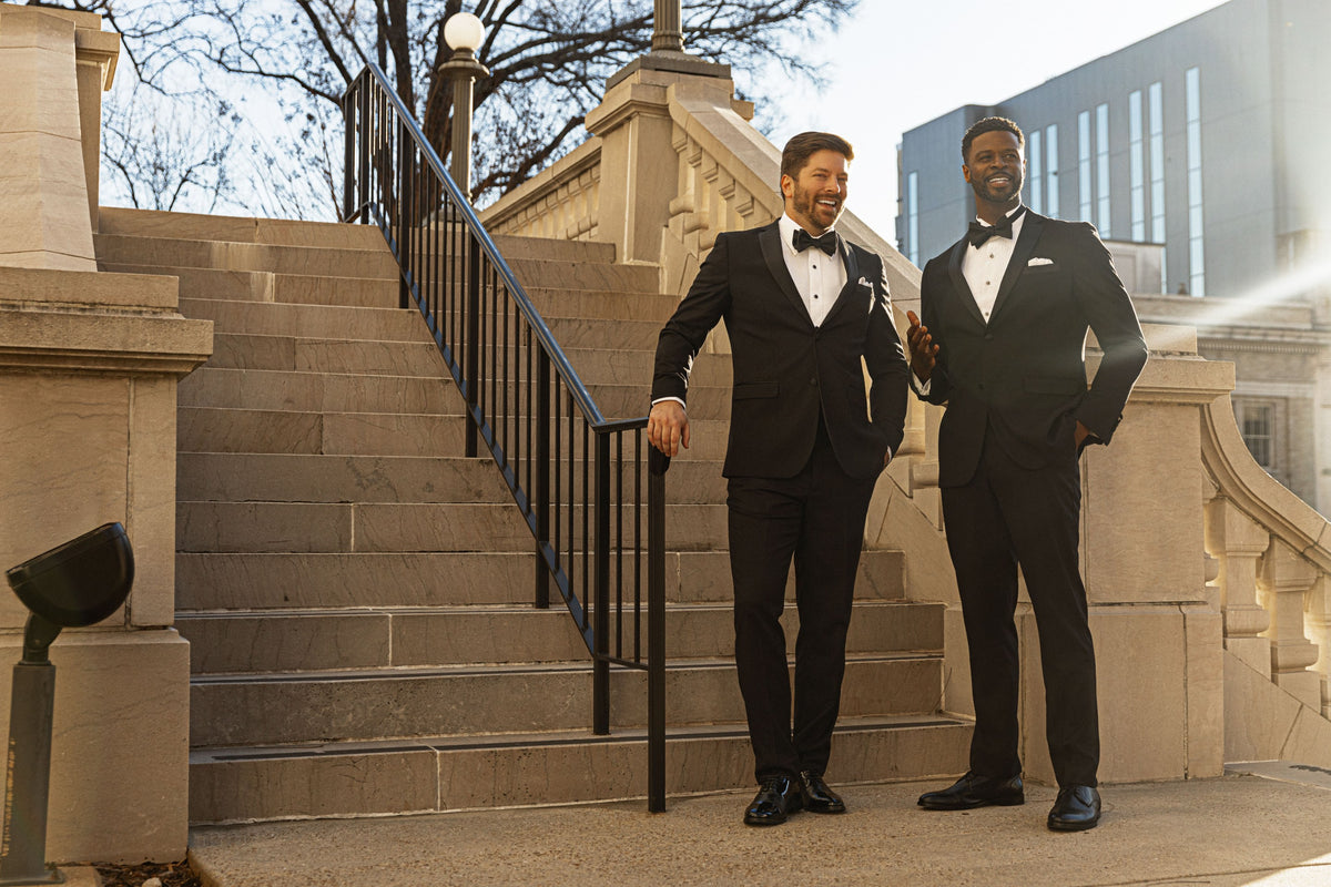 Two models in black tuxedos laughing together on stone courthouse steps