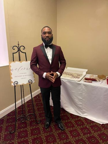 Chijioke O. in the Burgundy Peak Lapel Tuxedo beside a wedding welcome sign