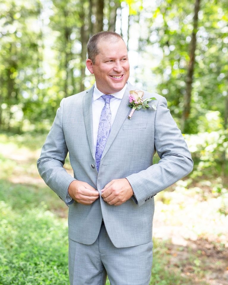 A smiling groom in a light gray suit and lavender tie buttoning his jacket outdoors