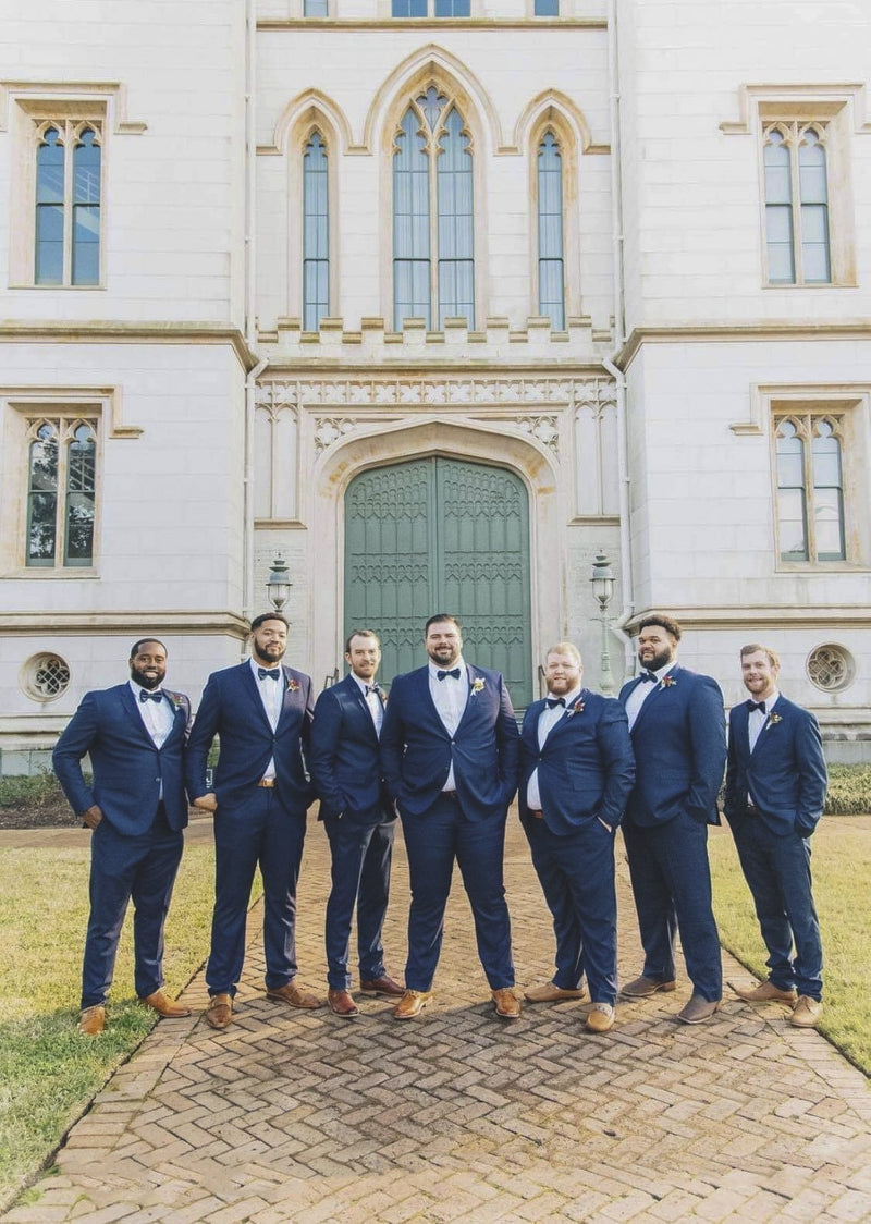 Seven groomsmen in navy suits and bow ties in front of a historic stone building